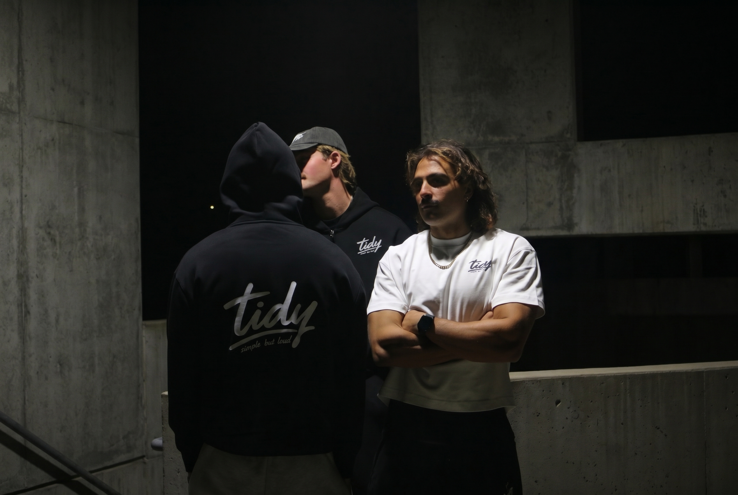 Two men wearing Tidy Design Australian streetwear — black OG heavyweight hoodie and white logo tee in an urban concrete setting